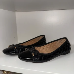 Svarta quiltade ballerinaskor Lady Glory - Snygga svarta ballerinaskor från Lady Glory i storlek 37. Skorna har quiltad yta, rund tå och en liten rosett framtill med guldfärgade detaljer. Insidan är mjukt vadderad för extra komfort och yttermaterialet är glansigt syntet.