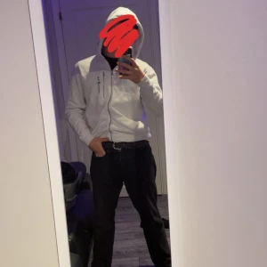 Vit hoodie från Sail Racing, storlek L - Vit hoodie från Sail Racing i storlek L. Tröjan har dragkedja framtill, huva med snörning och svart logga på bröstet. Tillverkad i mjukt bomullsmaterial och har ribbade muddar. Perfekt för chill dagar eller när du vill ha en clean look och den har ett litet hål på höger armen 