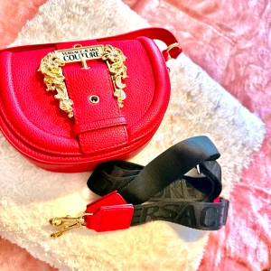 Röd axelväska från Versace Jeans Couture - Snygg röd axelväska från Versace Jeans Couture med rundad form och exklusiva guldfärgade detaljer framtill. Väskan är tillverkad i präglat skinn och har en bred svart axelrem med Versace-logga. Perfekt för dig som vill sticka ut med en lyxig accessoar.