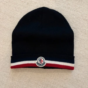 Svart Moncler mössa med logga - Svart mössa från Moncler med klassisk rund form och ribbad kant i marinblått, vitt och rött. Framtill sitter Monclers broderade logga. Mössan är stickad och tillverkad i Italien, perfekt för dig som gillar stilrena accessoarer med lyxig känsla.