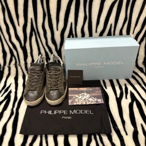 Philippe Model - Snygga svarta och grå sneakers från Philippe Model Paris med vita detaljer på hälen. Skorna har snörning, rund tå och är tillverkade i skinn och mocka. Klassisk låg modell med logga på sidan och slitstark gummisula. Og med följes dustbag, box & originalkorten. Minsta fundering hör av dig