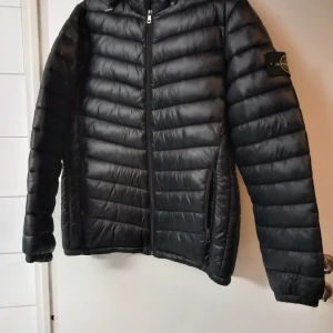 Svart lättviktsjacka med huva. - Svart lättviktsjacka med huva och logga på ärmen. Dragkedja framtill och två fickor. Storlekslappen är bortklippt, gissar på herr L i storlek, Mått: 70 cm längd, 65 cm ärm, 52 cm bredd. Köpt utomlands. Använd någon gång bara så fint skick.   