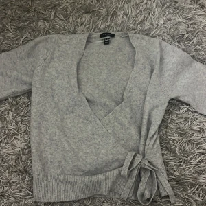 Grå omlottkofta med knyt från ASOS - Mysig grå omlottkofta från asos med ribbade muddar och snygg knytning i sidan. Koftan har långärmade ärmar och är tillverkad i ett mjukt stickat material som känns skönt mot huden. 