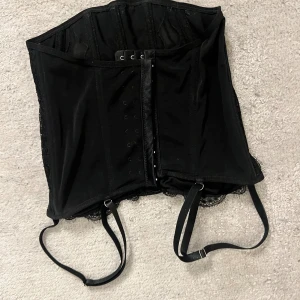 Svart korsett med spets och snörning - Säljer en svart korsett med snygga spetsdetaljer och justerbara remmar. Framsidan har hakar för stängning och baksidan är dekorerad med spetsmönster. Perfekt för dig som vill ha en edgy och stilren look.