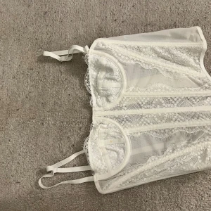 Vit spetskorsett med justerbara band - Säljer en vit korsett med transparent mesh och detaljerad spets. Toppen har justerbara axelband och hakknäppning i ryggen för perfekt passform. Spetsen har fina blommönster och ger en romantisk vibe. Perfekt för dig som vill ha något extra till outfiten.