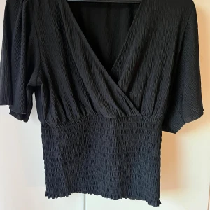 En svart söt topp  - En svart topp som är tightare vid midjan, i fint skick och knappt använd och har en liten omlott vid bröstet.