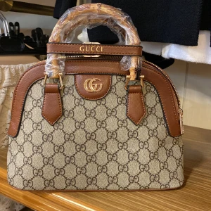 Gabriella handväska  - Snygg handväska  i beige och brunt. Väskan har bruna skinn-detaljer, guldfärgade metalldelar och bambuhandtag. Dragkedja upptill , Perfekt storlek för det viktigaste och riktigt lyxig vibe.