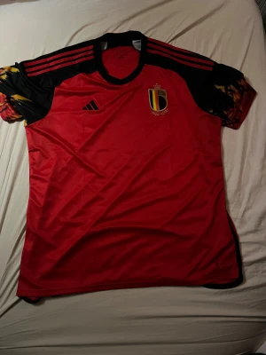 Belgien matchtröja Adidas 2XL - Säljer en officiell Belgien landslagströja från Adidas i storlek 2XL. Tröjan är röd med svarta och gula detaljer på ärmarna, har korta ärmar och broderat landslagsemblem på bröstet. Materialet är lätt polyester, perfekt för dig som gillar att samla på fotbollströjor. Skriv gärna till mig om ni har några frågor. Har aldrig haft på mig den