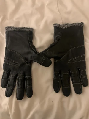 Svart skinnväst med pälsdetalj - Snygg svart väst från Warm Gloves med mjuk pälsdetalj längs kragen. Västen har markerade sömmar och dragkedja framtill för extra stilpoäng. Materialet är skinn och insidan har ett fluffigt foder som håller dig varm. Perfekt för dig som gillar en cool och modern look.