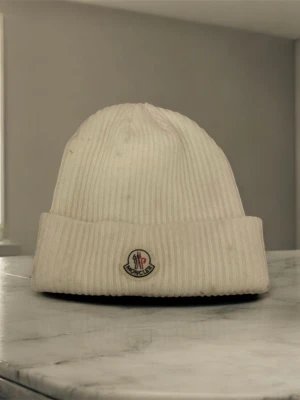 Vit ribbad mössa från Moncler - Snygg vit mössa från Moncler med ribbad struktur och uppvikt kant. Mössan har en klassisk Moncler-logga framtill och är stickad i mjukt material som håller dig varm. Perfekt för dig som vill ha en clean och stilren look under kalla dagar. Dessutom har mössan en liten svart fläck. Men det ändrar inte helheten på mössan.