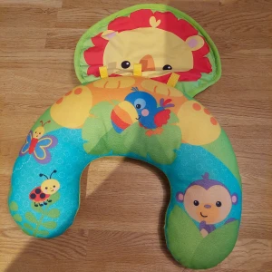 Färgglad djurmönstrad babyväst Fisher-Price - Säljer en superfin babykudde från Fisher-Price med färgglada djurmotiv som lejon, apa, fågel och fjäril. Västen har en U-formad design och är tillverkad i mjuk polyester. Perfekt för lek och mys med gulliga detaljer i grönt, blått, gult och rött.