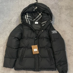Burberry pufferjacket  - En snygg pufferjacka från Burberry London England med stor patch på ärmen och klassiskt rutigt foder i huvan. Jackan har dragkedja, knappar och ribbade muddar vid ärmslut. Avtagbara ärmar så det går även att använda den som en väst. Perfekt för kalla dagar med sin tjocka och fluffiga design.