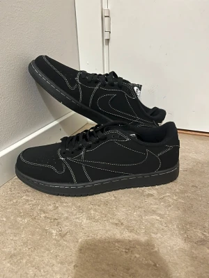 Nike Air Jordan 1 Low svart/vit - Svarta Nike Air Jordan 1 Low med vit kontrastsöm och broderade detaljer på hälen. Skorna har klassisk snörning, rund tå och platt sula. Materialet är mocka och syntet, och de har den ikoniska Air Jordan-loggan bak. Perfekta för dig som gillar stilrena sneakers.