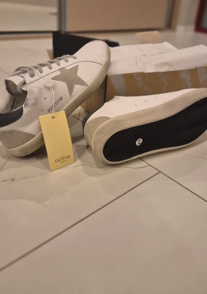 Golden Goose sneakers - Golden Goose sneakers i vitt skinn med grå mockadetaljer och en ikonisk stjärna på sidan. Skorna har grå snörning, rund tå och platt sula. Bakpartiet är svart och sulan är beige. Levereras med originalkartong och dustbag.