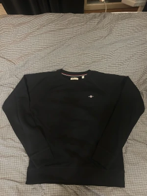 Svart sweatshirt från GANT - Svart sweatshirt från GANT med rund halsringning och diskret broderad logga på bröstet. Tröjan har ribbade muddar och en snygg detalj med röd och vit rand på insidan av kragen. Tröjan har alltid varit för liten för mig därför är den oanvänd vilket betyder den är precis som ny. Storlek 15y skulle jag säga sitter bäst på nån runt 170cm.