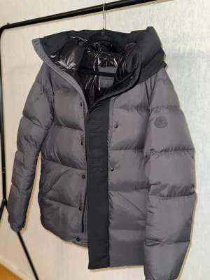 Moncler madeira - Säljer en svart pufferjacka från Moncler med stor huva och diskret logga på ärmen. Jackan har breda, quiltade sektioner, dragkedja och tryckknappar framtill samt innerfoder i glansigt material. Perfekt för kalla dagar och riktigt snygg streetstil. Helt ny jacka har aldrig används.