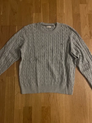 Grå kabelstickad tröja från GANT - Snygg grå kabelstickad tröja från GANT med rund hals och broderad logga på bröstet. Tröjan har ribbade muddar och är långärmad, perfekt för dig som gillar klassisk stil med en modern touch. Passar dig som vill ha en enkel men stilren look.