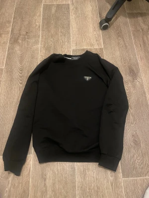 Svart sweatshirt från Prada - Svart sweatshirt från Prada med rund hals och långärmad design. Tröjan har en diskret logotyp på bröstet och ribbade muddar vid ärmslut och nederkant. Perfekt för dig som gillar stilrena och exklusiva plagg.