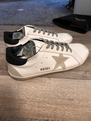 Golden Goose vita sneakers  - Golden Goose sneakers i vitt skinn med grå mockadetaljer och en ikonisk stjärna på sidan. Skorna har grå snörning, svart hälparti och platt sula. Snygg och stilren design som sticker ut med den klassiska GGDB-loggan och kontrastfärger.