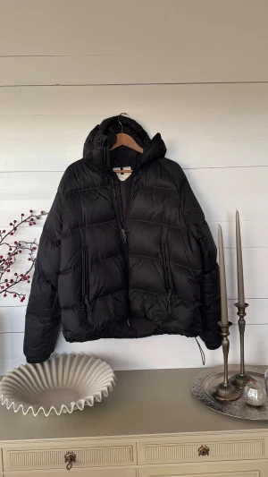 Svart pufferjacka från Arket - Svart pufferjacka från Arket med huva och två stora fickor med dragkedja framtill. Jackan har snörning i nederkant och är quiltad för extra värme. Perfekt för kalla dagar. Endast använd ett fåtal gånger. Nypris: 2790kr 