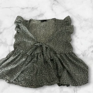 Grön blommig blus med volang - Supersöt grön blus med ljusa blommönster, volangdetaljer vid axlarna och knyt framtill. Blusen har en v-ringad hals och är kortärmad, perfekt för att ge din outfit en romantisk vibe.