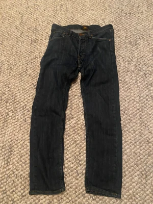 Mörkblå jeans från Lee - Snygga mörkblå jeans från Lee med klassisk femficksdesign och brunt Lee-märke på bakfickan. Jeansen har raka ben och kontrastsömmar i gult, vilket ger en tidlös look. Materialet är slitstarkt denim i bomull.