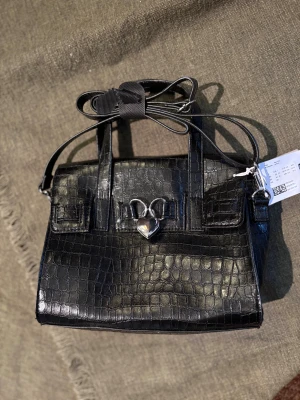 Svart handväska med hjärtdetalj från Monki NY med lapp - Stilren svart handväska från Monki i croco-mönstrad syntet med silverfärgat hjärtlås framtill. Väskan har både handtag och avtagbar axelrem, perfekt för dig som gillar edgy accessoarer med en söt twist.