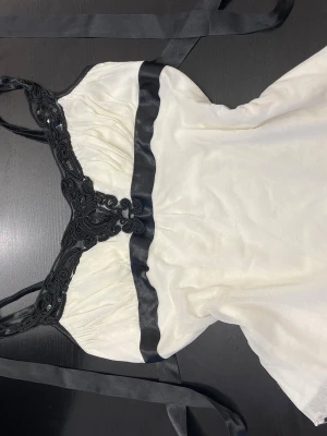 Vit och svart korsett med paljetter - Säljer en snygg korsett-topp i vit och svart med detaljerad broderad spets och svarta paljetter vid bysten. Toppen har svarta satinband som kan knytas i ryggen och smala axelband. Materialet känns mjukt och har en lyxig känsla, perfekt för att sticka ut.