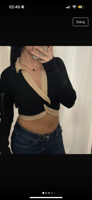 Svart croppad omlottopp från Stories - Säljer en svart croppad omlottopp från Stories i ribbad stickad kvalitet. Toppen har långa ärmar och snygga beigea detaljer längs krage och nederkant. Perfekt för dig som gillar trendiga och unika plagg med twistad front.