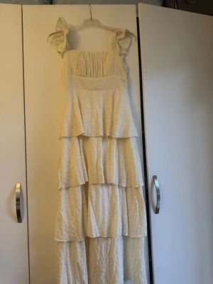 Ljus volangklänning med axelband - Superfin långklänning i ljus beige med flera volanger och smockad överdel. Axelbanden har volangdetaljer som ger en romantisk känsla. Klänningen är tillverkad i ett lätt, krinklat tyg som känns luftigt och somrigt.