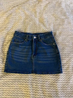 Mörkblå jeanskjol med fickor - Snygg mörkblå jeanskjol i klassisk modell med fem fickor, bälteshällor och knapp framtill. Kjolen är kort och har en rak passform som ger en cool och tidlös look. Perfekt att matcha med en t-shirt eller hoodie för en avslappnad stil.