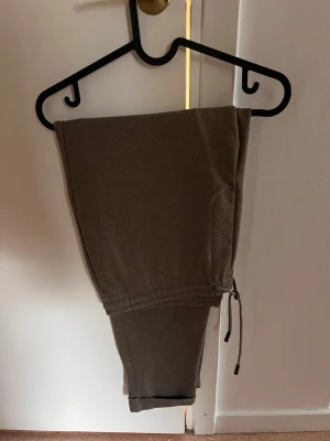 Bruna kostymbyxor med snörning - Säljer ett par bruna kostymbyxor med  bra passform. Byxorna har muddar vid bensluten och är tillverkade i ett mjukt bomullsliknande material. Perfekta för chill dagar eller när du vill vara bekväm men ändå se snygg ut.