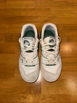 New Balance 550 vita och gröna sneakers, stl 41.5 - Säljer ett par New Balance 550 sneakers i vitt och mörkgrönt med klassisk retrodesign. Skorna har snörning, platt sula och rund tå. Ovandelen är i skinn och syntet med gröna detaljer och New Balance-logga på sidan. Perfekt för dig som gillar streetwear och sportig stil. Storlek 41.5