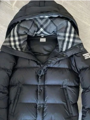 Svart pufferjacka från Burberry - Säljer en svart pufferjacka från Burberry med avtagbara ärmar så den kan användas som väst. Jackan har rutigt foder i huvan, dragkedja och knappar framtill samt fickor med dragkedja. Klassisk Burberry-logga på ärmen och stilren design.