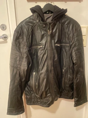 Svart skinnjacka med huva från Molltan - Svart skinnjacka från Molltan med cool huva i sweatshirtmaterial. Jackan har flera dragkedjeförsedda fickor fram, quiltade detaljer på axlar och ärmar samt långa ärmar med dragkedja vid mudden. Perfekt för dig som gillar streetstyle och vill ha en edgy look.