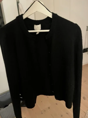 Svart kofta från H&M, storlek M - Svart, klassisk kofta från H&M i storlek M. Koftan har rund hals, långa ärmar och knappar framtill. Materialet är mjukt och stickat, perfekt för lager-på-lager under kyliga dagar. Enkel och stilren design som funkar till många olika looks.