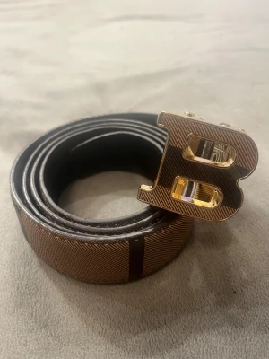 Balmain Paris bälte - 🔥 Balmain Paris Bälte – Ikoniskt Guldspänne 🔥 Säljer ett exklusivt Balmain Paris-bälte med det klassiska B-spännet i guld, en riktig statement-accessoar för dig som vill sticka ut med stil. Det eleganta bruna bandet med fin textur ger en premiumkänsla som Balmain är kända för.  ✨ Detaljer: 	•	Äkta Balmain Paris 	•	Ikoniskt guldigt B-spänne 	•	Lyxig brun rem med exklusivt mönster 	•	Sitter perfekt till jeans, kostym och streetwear 	•	I mycket fint skick – ser knappt använt ut  Det här bältet höje