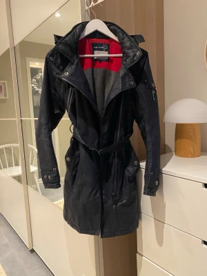 Jacka  - Marinblå jacka från Pelle P med huva, bälte i midjan och snygga detaljer som knappar och dragkedjor. Insidan har röd och grå kontrastfärg. Jackan har långa ärmar, flera fickor och logotyp på både insidan och ryggen. Perfekt för dig som gillar stilren och funktionell design. Storlek S.