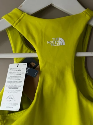 Neongul topp från The North Face - Säljer en neongul linnetopp från The North Face med sportig racerback-design. Toppen är ärmlös och har en tight passform, perfekt för träning. Materialet är stretchigt och består av syntetfiber med Lycra för extra komfort. Logga tryckt i vitt på ryggen.