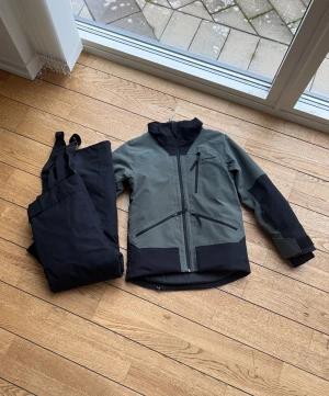 Grön och svart vindjacka från Everest - Säljer en grön och svart vindjacka från Everest med huva och flera praktiska fickor med dragkedja. Jackan har lång ärm och är tillverkad i slitstark syntet, perfekt för utomhusaktiviteter. Snygg, sportig design med kontrastpaneler och justerbara muddar.