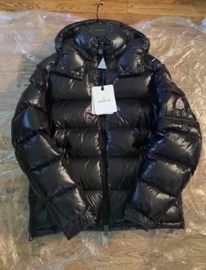 Svart Moncler pufferjacka med huva - Säljer en svart, glansig pufferjacka från Moncler med huva och dragkedja framtill. Jackan har klassisk quiltad design, Moncler-logga på ärmen och innerficka med dragkedja. Perfekt för kalla dagar när du vill hålla stilen.