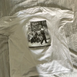 Vit t-shirt med Birra Peroni tryck från Bvaldi - Vit t-shirt från Bvaldi med ett stort svartvitt foto-tryck på framsidan där det står 'Birra Peroni'. Klassisk rund hals och korta ärmar. Materialet känns som mjuk bomull och passformen är normal. Perfekt för dig som gillar retro vibes och coola prints.