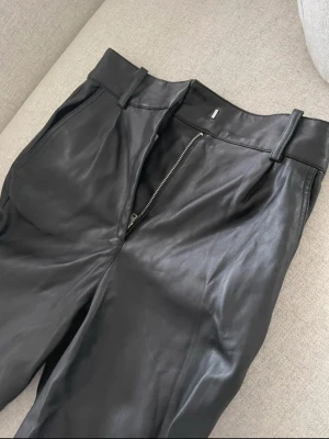 Svarta skinnbyxor strl 36 - Snygga svarta byxor i skinnimitation med hög midja och raka ben. Byxorna har bälteshällor, sidofickor och dragkedja framtill. Mjuk på insidan. Inga fickor bak. Snäppet kortare i benen. Ankellånga liksom
