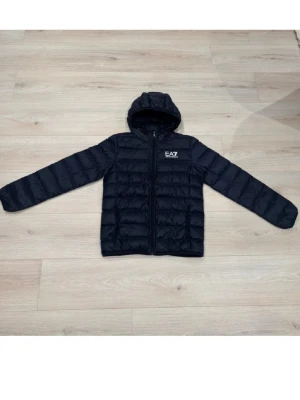 Svart dunväst med huva från EA7 - Svart dunväst från EA7 med quiltad design och huva. Västen har dragkedja framtill och EA7-logga på bröstet. Perfekt för dig som gillar stilren och sportig look. Passformen är normal och materialet är lätt och bekvämt.