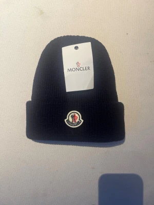 Mörkblå stickad mössa från Moncler - Snygg mörkblå stickad mössa från Moncler med klassisk Moncler-logga framtill. Mössan har en ribbad struktur och uppvikt kant som ger en cool och tidlös look. Perfekt för kalla dagar och passar till streetwear eller vinteroutfits.