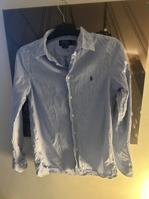 Blå skjorta från Polo Ralph Lauren - Snygg ljusblå skjorta från Polo Ralph Lauren i slim fit-modell. Skjortan har klassisk krage, långa ärmar och knappar framtill. Broderad logga på bröstet. Perfekt för dig som gillar stilrena och tidlösa plagg.