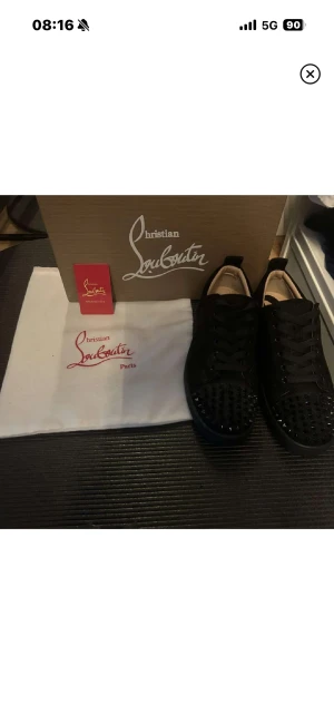 Christian Louboutin svarta sneakers - Svarta sneakers från Christian Louboutin med ikoniska röda sulor och svarta nitar på tån. Skorna har snörning och är tillverkade i mocka med platt sula och rund tå. Snygg och edgy design som sticker ut med detaljerna och den lyxiga känslan.