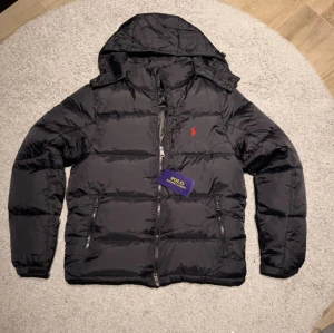 jacka Polo Ralph Lauren - Svart pufferjacka från Polo Ralph Lauren med huva och dragkedja framtill. perfekt för kyliga dagar, för ett bra pris. Kom dm vid frågor😇