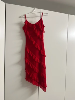 Röd volangklänning med smala axelband - Säljer en röd långklänning med smala axelband och flera diagonala volanger som ger en riktigt snygg och livlig look. Klänningen har asymmetrisk nederkant och är gjord i ett lätt, flödande material som känns skönt mot huden.