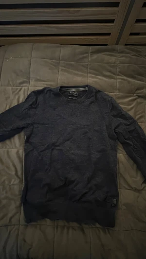 Mörkblå stickad tröja East West - Mörkblå stickad tröja från East West med rund hals och ribbade muddar. Snygg detalj med ribbad V-form vid halsen och en liten logotyp-patch nertill. Perfekt för dig som gillar enkel och stilren design.